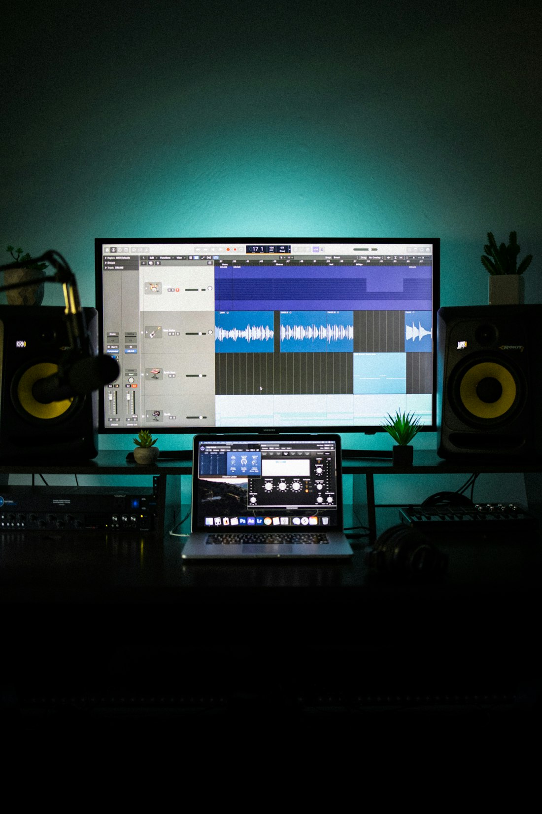 A dark home studio with a DAW session open on the monitor and studio speakers glowing teal